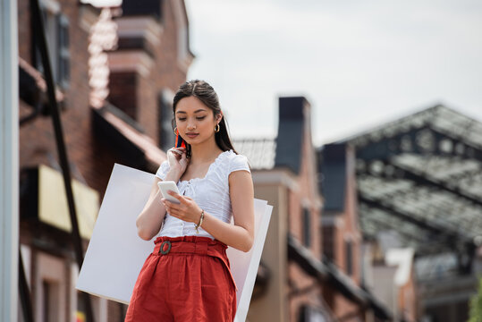 Young Asian Woman With Shopping Bag Messaging On Mobile Phone Outdoors