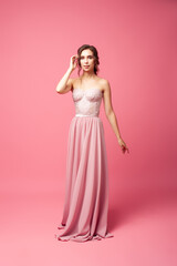 A young woman in a pink dress stands on an isolated background. A beautiful young woman in a long silk dress stands on a pink background. 