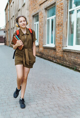 Obraz premium Teen schoolgirl on the way to the school. She is listening to music on headphones and holding a book. Education and back to school concept