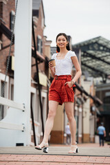 Obraz premium joyful asian woman holding coffee to go while walking on urban street with hand in pocket