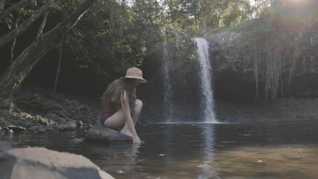 Wide shot of a young woman in swimwear sitting on a rock in front of a waterfall and playing with the water