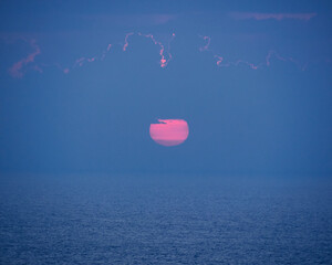 Fototapeta premium sunset, a magenta pink sun setting over the horizon with a calm sea and a sky with few clouds
