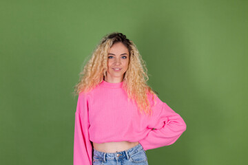 Young blonde woman with long curly hair in pink sweater on green background positive smiling look to camera