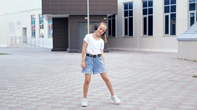 Schoolgirl in earphones dances. Young teenager in t-shirt denim shorts and black wireless device listens to music in school yard against building