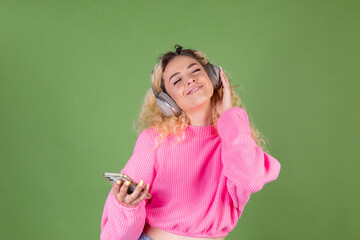 Young blonde woman with long curly hair in pink sweater on green background happy positive enjoying favorite music in wireless headphones