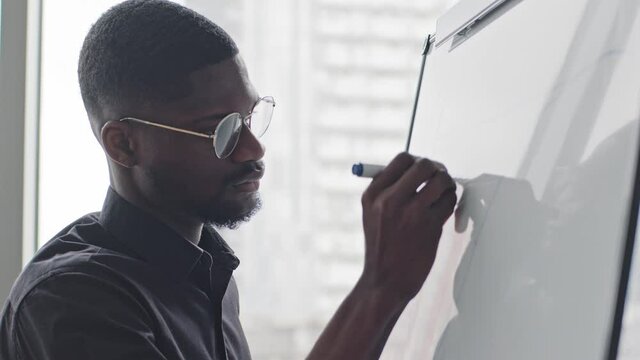 Concentrated african american afro black male manager in glasses writing notes on white board. Focused professional business man preparing for project presentation, drawing charts on office flipchart
