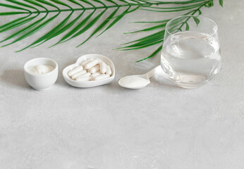 Collagen protein powder, pills and a glass of water on a gray background with green leaves. Supplement for bone and skin health. Healthy lifestyle. Space for text.
