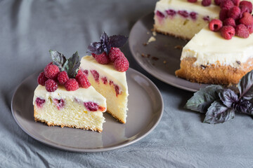 Raspberry cheesecake slices with fresh raspberries and bazil leaves on a grey plate, whole cheesecake in background. Selective focus