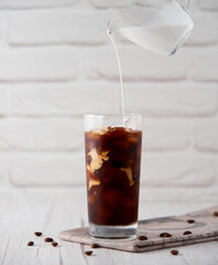 Iced coffee in a tall glass with cream or milk poured over and coffee beans and straws on light background. Cold tasty summer refreshment beverage concept. Selective focus, copyspace.