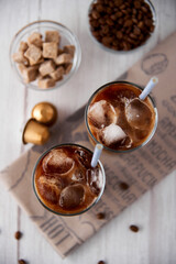Iced coffee latte in a tall glass with cream and beans, golden capsules, straws on light background. Cold tasty summer refreshment beverage concept. Selective focus, copyspace. Flat lay, top view.