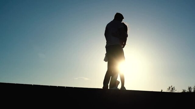 Silhouette of loving young couple in love dancing and kissing at sunset on background of cloudless sky. Wide shot of romantic happy couple hugging outdoor. Shooting in slow motion