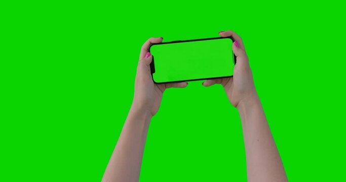 Female woman hands with pink manicure playing games on the smartphone on green screen chroma key background. Mobile phone mock-up for your product. iPhone 12 Pro Max model in horizontal orientation.