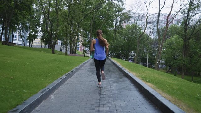 Slow motion woman with ponytail wearing sportswear jogging on wet walkway in park. Following shot fit female training outside after rain. Concept of fitness