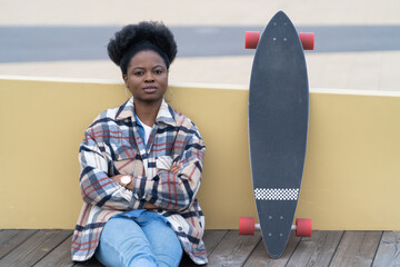 Young african american woman relaxing after skateboarding on city street near river sit calm recreate. Black millennial female alone with longboard ponder. Urban lifestyle and leisure activity concept