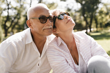 Mustachioed man in black sunglasses and white cool outfit posing with blonde smiling woman in pink...