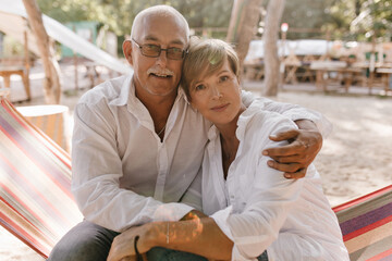 Old man with gray mustache and glasses in shirt looking into camera and hugging blonde short haired woman in white clothes on beach..