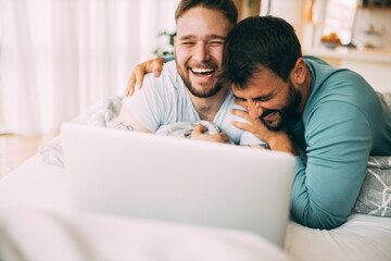 Happy gay couple in love lying in their bed and using a laptop. They are laughing and hugging. Love celebration concept.