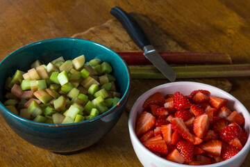fresh strawberries and rhubarb cut to pieces in bowls on the surface of a table together with a knife, a cutting board and two rhubarb stalks