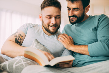 Happy gay couple sitting at home in the morning and reading an exciting book. It takes a little to...