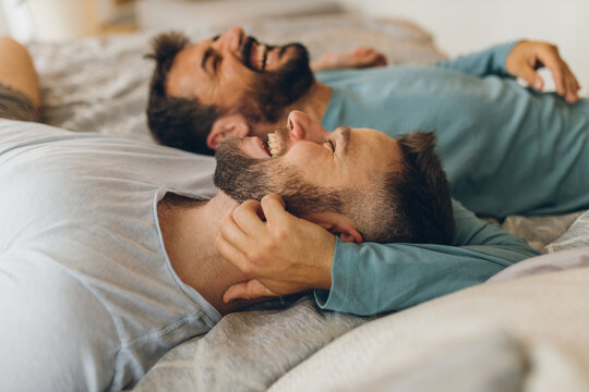 Happy gay couple lying down on the bed at home, hugging and flirting