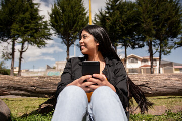 young happy latina woman with long hair sitting on the grass using her cell phone, concept of happiness