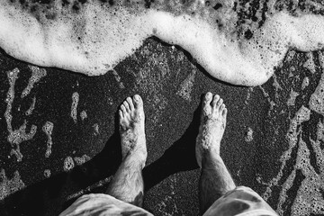 Man feet in the sand in front a foamy wave (in black and white)