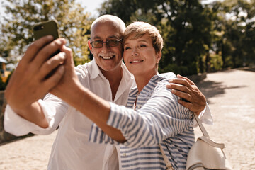 Wonderful woman with short blonde hairstyle in blue striped outfit making photo with grey haired man with mustache in white clothes in park..