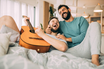 Happy gay couple in love having romantic times at home. Man is playing guitar to his beloved...