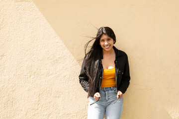 young happy latin woman with long hair leaning against a yellow wall, concept of happiness