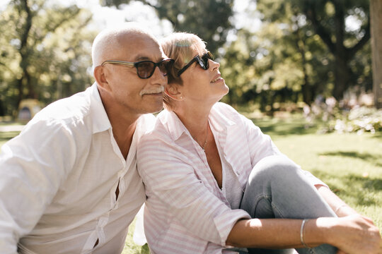 Grey haired man with mustache in sunglasses and light shirt smiling and sitting on grass with woman with short hair in pink blouse outdoor..