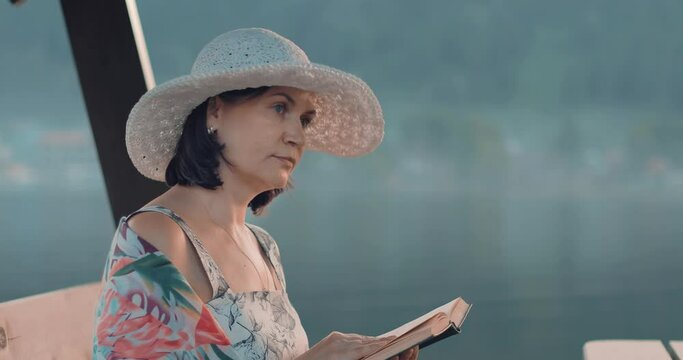 A beautiful girl in a hat sits in the gazebo by the lake in the evening and reads a book. The content of the book worries the girl and she sighs, reliving what she read. The girl reads a book in natur
