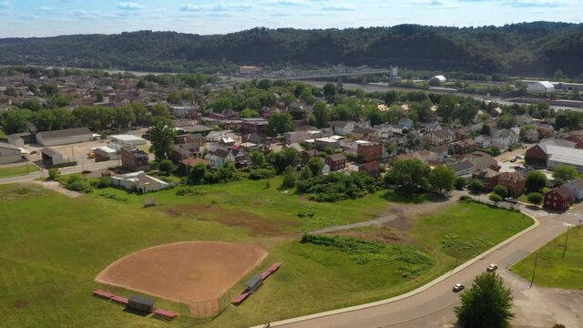 A slowly moving forward aerial view of a small Pennsylvania river town. A baseball sandlot field in the foreground and the Ambridge Bridge over the Ohio River in the distance.  	