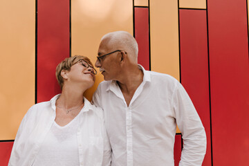 Wonderful woman with blonde short hairstyle in white clothes and eyeglasses posing with grey haired old man in light shirt on orange and red backdrop..