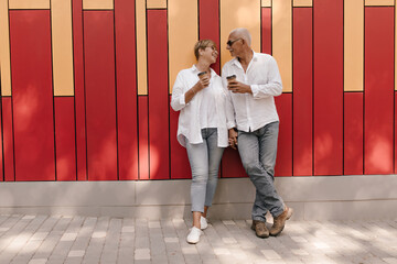 Smiling woman in white blouse and jeans posing with cup of tea and holding hands with man in light shirt on red and orange backdrop..