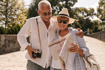 Cheerful man with grey hair in light shirt and jeans with camera smiling and looking at map with blonde lady in hat and blue outfit in park..