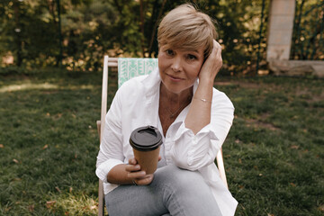 Charming woman with short hairstyle in white shirt and jeans holding cup of coffee, looking into camera and sitting in park..