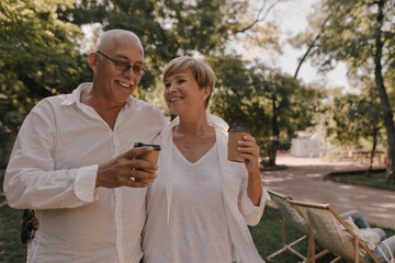 Fototapeta premium Grey haired man with eyeglasses in white shirt smiling and posing with cap of tea and stylish woman in light outfit outdoor..