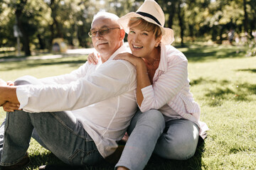 Fashionable lady with short stylish hairstyle in pink blouse, hat and jeans laughing and sitting on grass with man in light shirt in park..