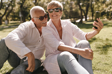Happy woman with blonde hair in pink clothes and sunglasses smiling, sitting on grass and making photo with grey haired man in white shirt in park..