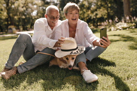 Positive lady with blond hair in striped shirt and jeans making photo with husband in eyeglasses and corgi in hat on outdoor..