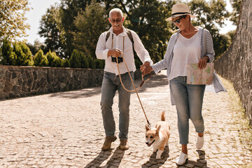 Wonderful woman in hat, white t-shirt, striped blouse and jeans holding card and hand grey haired man in light clothes with dog in park..