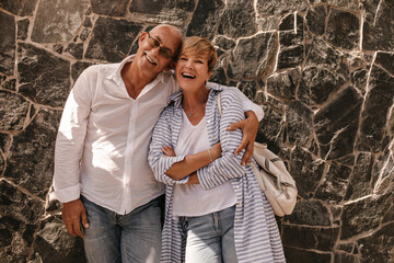 Cheerful woman with blonde hairstyle in blue shirt and jeans laughing and hugging with mustachioed man in white shirt on background of wall..