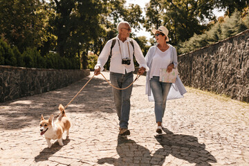 Trendy woman in hat and blue blouse walking and looking grey haired man in white long sleeve shirt with camera and corgi in park..