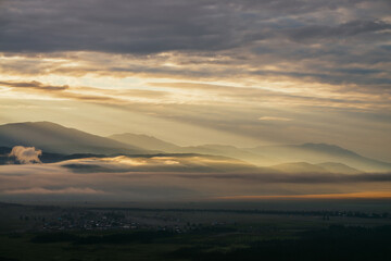 Scenic mountain landscape with golden low clouds above village among mountains silhouettes under dawn cloudy sky. Atmospheric alpine scenery of countryside in low clouds in sundown illuminating color.