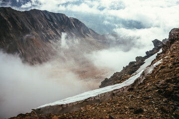 View from precipice edge with sharp rocks and snow cornice to mountain valley in low clouds. Wonderful mountain scenery with low clouds in highland valley. Scenic alpine landscape with rocky mountains