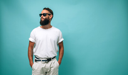 Fototapeta premium Portrait of handsome, young, stunning, perfect bearded guy wearing a white blank t-shirt stands against turquoise background. T-shirt template.