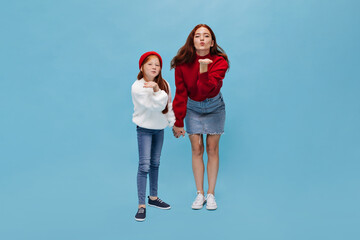 Stylish two sisters in stylish outfit blows kisses on blue background. Red haired girl in white...