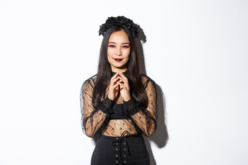 Devious young asian woman in gothic lace dress smiling pleased and steeple fingers. Witch prepare...