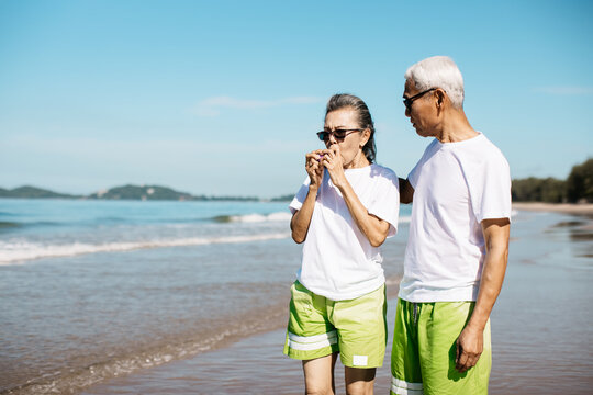 Senior Wife Having Shortness Of Breath From Asthma While Strolling Along The Beach With Her Husband. Using A Nebulizer And Inhaler. Older Man Take Care And Support Wife, Health Problem And Insurance.