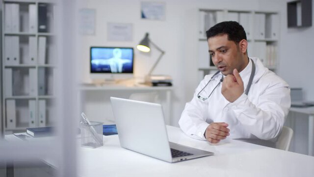 Middle Eastern medical specialist wearing white coat sitting in his office in front of laptop working with patient on video call during lockdown
